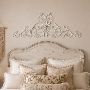 May include: A white metal wall art piece featuring intricate scrollwork and floral motifs mounted above a bed. The decorative element has a symmetrical design with curved lines and small leaf details, providing a focal point for the bedroom wall.