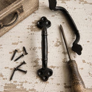 May include: Two black cast iron drawer pulls with decorative ends, each featuring three screw holes. The set includes four black metal screws and a flathead screwdriver with a wooden handle, all arranged near a small wooden box.