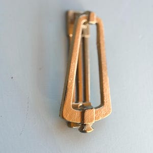 May include: A brass-colored metal door knocker featuring a tall, trapezoidal frame with a central vertical bar. The design includes a small, rounded knob at the bottom and a hinged mounting plate at the top, showcasing a textured, metallic finish.