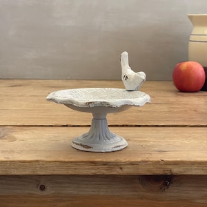 May include: A small, white, pedestal-style dish featuring a sculpted bird perched on the rim. The dish has a distressed finish with visible brown undertones, a scalloped edge, and a shallow bowl with a subtle floral pattern embossed on the interior.