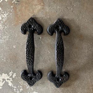 May include: Two black, textured metal door pulls. Each pull has a handle with a decorative top and bottom, and a hammered appearance. The pulls are designed for easy gripping and are likely for a cabinet or door.