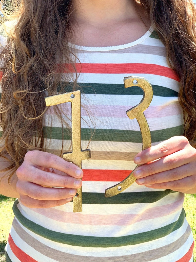 May include: Gold-painted wooden numbers '1' and '3' held in front of a striped shirt. The numbers are cut from wood and have a metallic gold finish. The numbers are likely for a house number or a birthday decoration.
