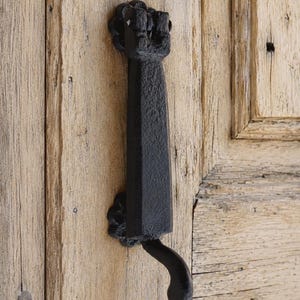 May include: A black cast iron door knocker with a textured finish, featuring a vertical rectangular body and a curved handle at the bottom. The knocker is mounted to a light-colored wooden door with visible grain and a panel design.