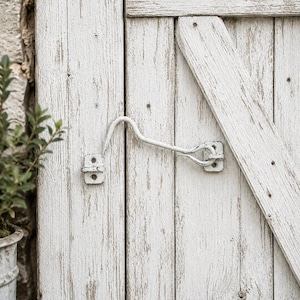 May include: A white metal cabin hook latch mounted on a weathered white wooden door with vertical planks and a diagonal brace. The hardware features a curved hook that secures into a small eye plate, showing signs of paint wear and texture.