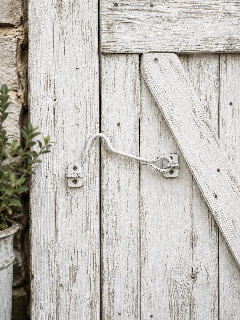 May include: A white metal cabin hook latch mounted on a weathered white wooden door with vertical planks and a diagonal brace. The hardware features a curved hook that secures into a small eye plate, showing signs of paint wear and texture.