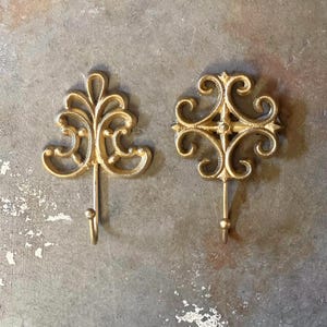 May include: Two gold-colored metal hooks with decorative scrollwork designs. The hook on the left has a more elaborate design with a floral motif. The hook on the right has a more geometric design with a cross shape.