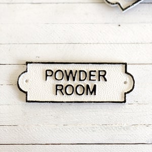 Puede incluir: Un letrero de hierro fundido blanco con letras negras que dice "POWDER ROOM".