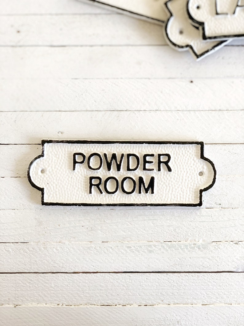 Puede incluir: Un letrero de hierro fundido blanco con letras negras que dice "POWDER ROOM".