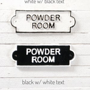 Puede incluir: Dos letreros de hierro fundido que dicen "Powder Room" en blanco y negro. El letrero superior es blanco con texto negro y el letrero inferior es negro con texto blanco.