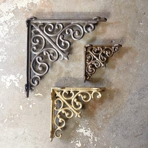 May include: Three decorative metal shelf brackets with a scrolled design. The largest bracket is black, the medium bracket is gold, and the smallest bracket is gold.
