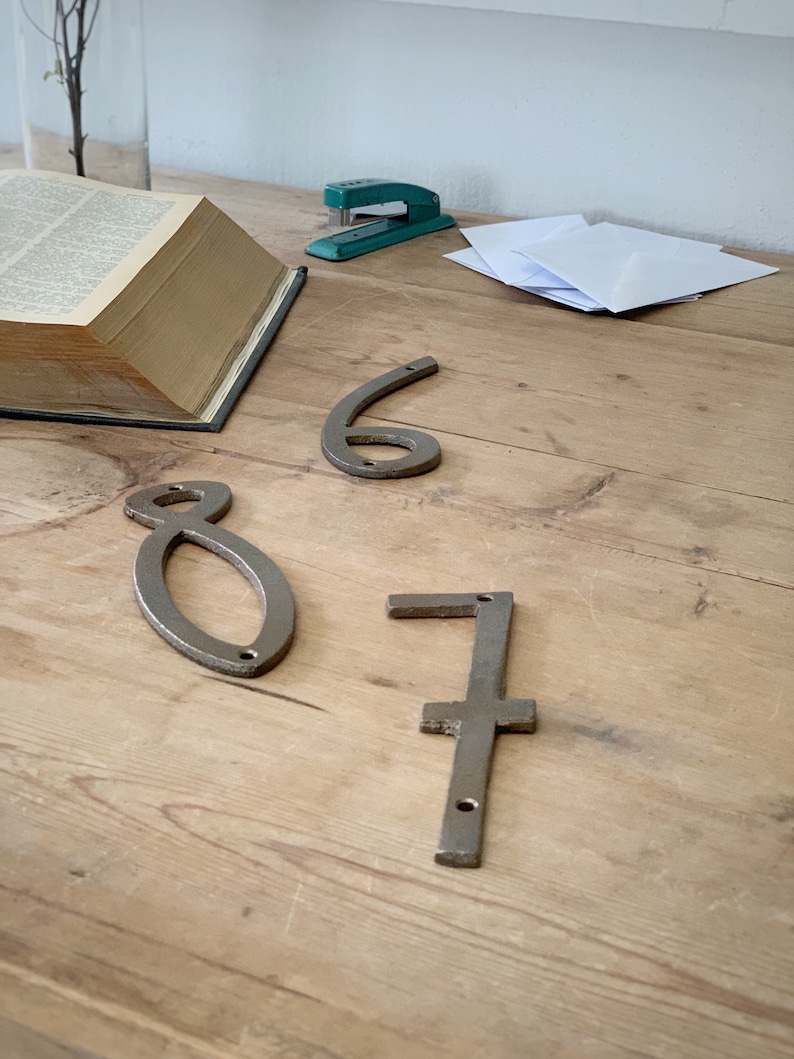 May include: Three metal house numbers, 6, 8, and 7, in a rustic, weathered finish, are displayed on a wooden surface. A green stapler, a stack of white envelopes, and an open book are also visible.