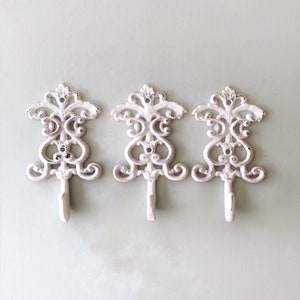 May include: Three white cast iron hooks with a decorative scroll design. The hooks are mounted on a light green wall.