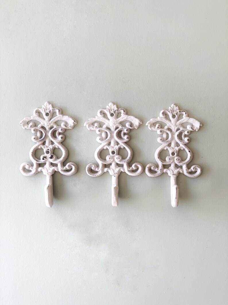 May include: Three white cast iron hooks with a decorative scroll design. The hooks are mounted on a light green wall.