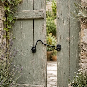 May include: A weathered sage green wooden garden gate featuring vertical planks and horizontal support beams. A black metal cabin hook latch is secured across the center of the two gate panels, holding them in a slightly open position.