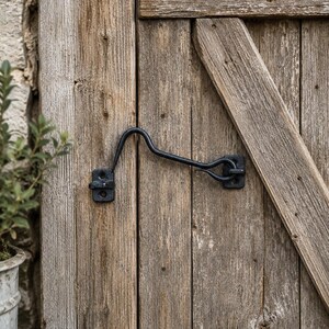 May include: A black metal cabin hook latch mounted on a weathered wooden door. The hardware consists of a curved hook arm and two rectangular mounting plates, each secured with two screws, designed to hold the door in a fixed position.