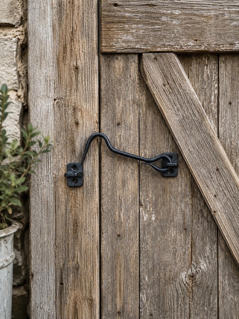 May include: A black metal cabin hook latch mounted on a weathered wooden door. The hardware consists of a curved hook arm and two rectangular mounting plates, each secured with two screws, designed to hold the door in a fixed position.