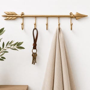 May include: A gold-colored metal wall hook rack shaped like an arrow with five individual hooks. A brown leather keychain with metal keys hangs from the second hook, and a beige fabric cloth hangs from the fourth hook on the arrow-shaped rack.