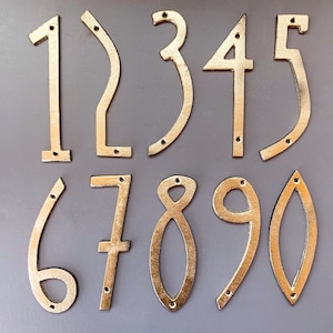 May include: A collection of ten gold-colored metal house numbers, ranging from 0 to 9. Each number has a unique shape and includes pre-drilled holes for mounting. The numbers are displayed against a gray background.