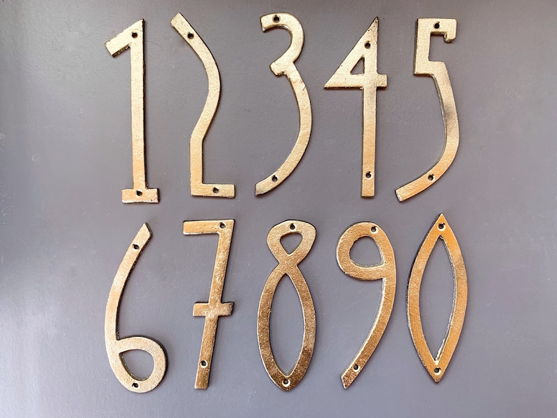 May include: A collection of ten gold-colored metal house numbers, ranging from 0 to 9. Each number has a unique shape and includes pre-drilled holes for mounting. The numbers are displayed against a gray background.