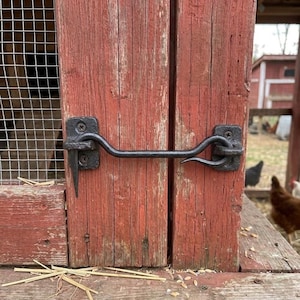 May include: A black metal cabin hook latch mounted on weathered red painted wooden boards. The hardware consists of a horizontal rod with a curved hook end that secures into a square plate, featuring visible screws and a textured dark finish.