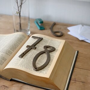 May include: An open book with the numbers 7 and 8 in a rustic metal finish resting on the pages. A clear glass vase with a plant and a teal stapler are in the background.