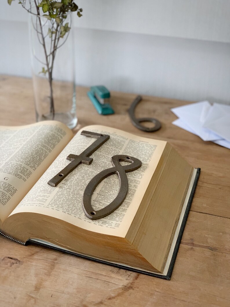 May include: An open book with the numbers 7 and 8 in a rustic metal finish resting on the pages. A clear glass vase with a plant and a teal stapler are in the background.