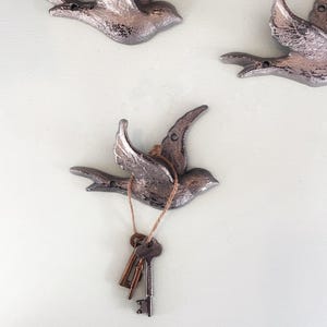 May include: Three silver-colored metal bird wall hooks. One bird holds a string with three keys attached. The birds are mounted on a light green wall. The hooks are decorative and functional.