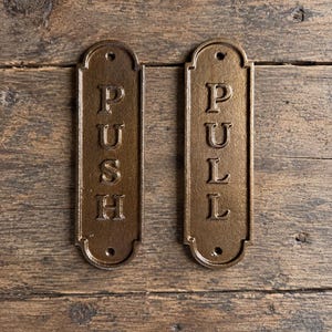 May include: Two rectangular metal door plates with rounded ends, finished in a dark bronze tone. One plate features the word PUSH in raised capital letters, and the other features the word PULL in raised capital letters. Each plate has two screw holes.