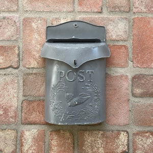 May include: A gray metal mailbox with a decorative floral design and a bird. The mailbox has a slot for mail and says "POST" on the front.