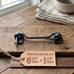 May include: A forged black iron hook latch with a rustic finish. A tan paper tag attached by twine displays the text: FORGED BLACK IRON HOOK LATCH, 6 1/2" WIDTH x 1 1/4" HEIGHT. The latch is positioned above a wooden ruler showing imperial measurements.