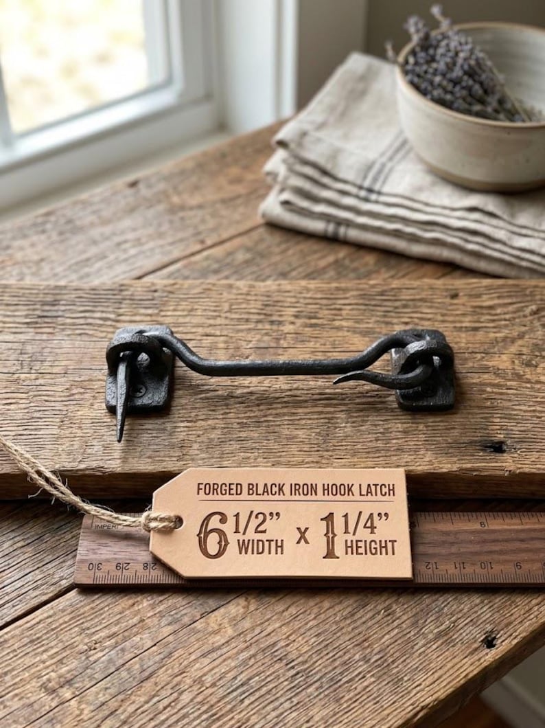 May include: A forged black iron hook latch with a rustic finish. A tan paper tag attached by twine displays the text: FORGED BLACK IRON HOOK LATCH, 6 1/2" WIDTH x 1 1/4" HEIGHT. The latch is positioned above a wooden ruler showing imperial measurements.