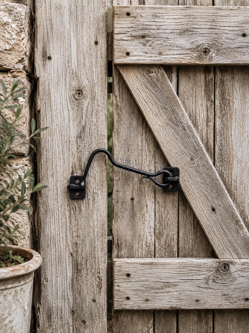 May include: A black metal cabin hook latch installed on a weathered wooden gate. The latch features a curved hook arm that secures into a mounting plate, both attached with visible screws to the vertical planks of the gray, textured wood door.
