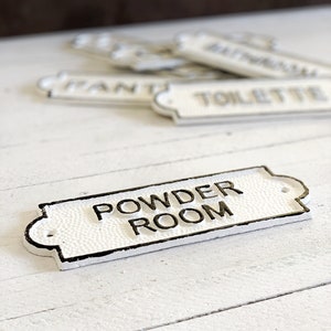 Puede incluir: Un letrero de hierro fundido blanco con letras negras que dice "Powder Room".