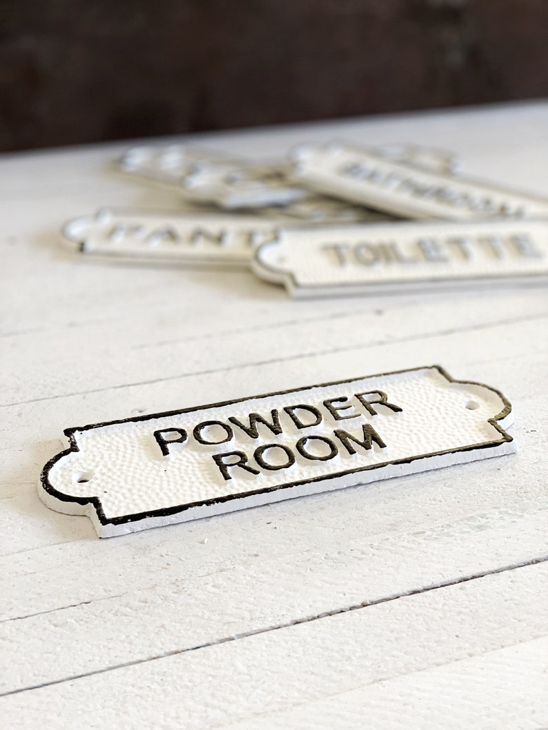 Puede incluir: Un letrero de hierro fundido blanco con letras negras que dice "Powder Room".