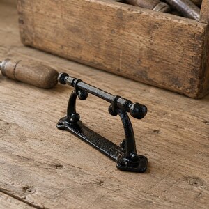 May include: A black cast iron handle featuring a horizontal bar supported by two curved uprights, each ending in a flat rectangular base with screw holes. The hardware has a textured, matte finish and a simple, functional design for mounting.