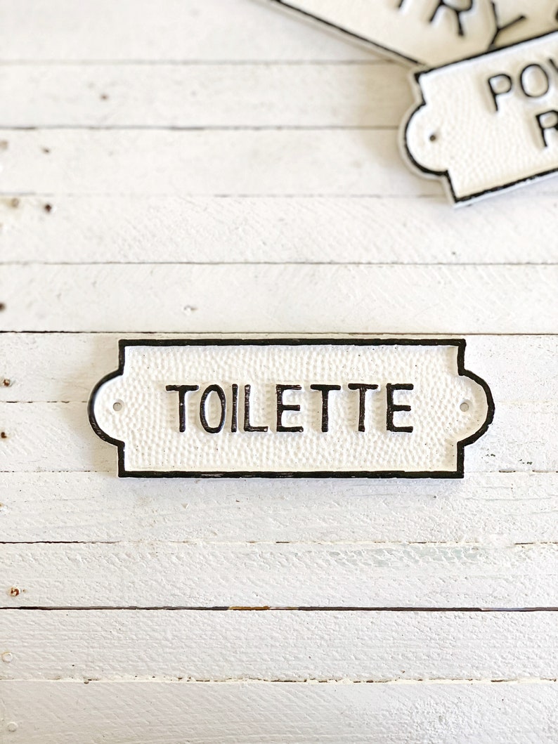 Puede incluir: Un letrero de hierro fundido blanco con letras negras que dice "TOILETTE".