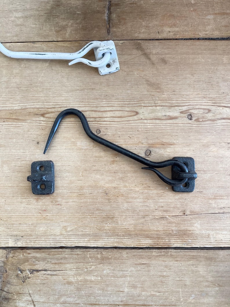 May include: A black metal cabin hook and eye latch set featuring a curved hook and a rectangular mounting plate with two screw holes, positioned alongside a separate white metal cabin hook and eye latch set of the same design.