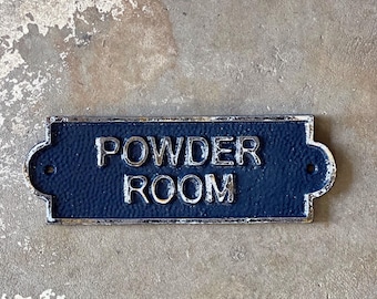 May include: A black cast iron sign with silver lettering that reads "POWDER ROOM". The sign has a distressed look and is mounted on a gray concrete wall.