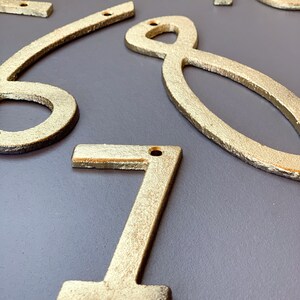 May include: Close-up of several gold-colored metal house numbers. The numbers are in various styles and sizes, with a textured finish. Each number has a small hole for mounting. The numbers are arranged on a gray surface.