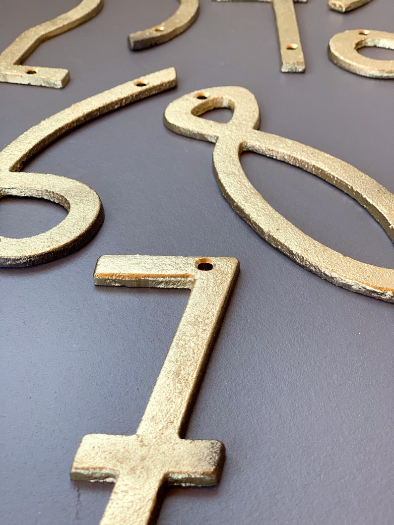 May include: Close-up of several gold-colored metal house numbers. The numbers are in various styles and sizes, with a textured finish. Each number has a small hole for mounting. The numbers are arranged on a gray surface.