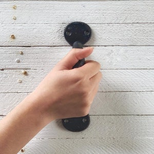 May include: Black metal door handle with a rounded shape. The handle is attached to a white wooden surface.