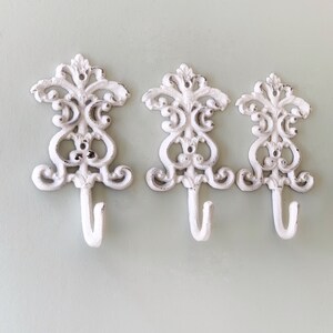 May include: Three white cast iron hooks with a decorative scroll design. The hooks are mounted on a light green wall.