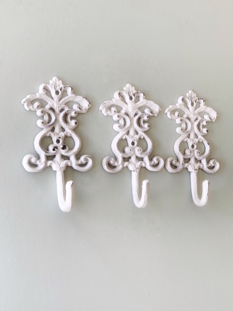 May include: Three white cast iron hooks with a decorative scroll design. The hooks are mounted on a light green wall.