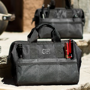 May include: Two black canvas tool bags with a zipper closure and multiple pockets. The bag on the left has the initials 'CR' embroidered on the front. The bag on the right has a red multi-tool sticking out of the side pocket.