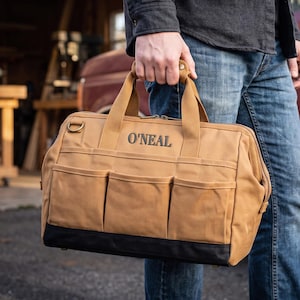 May include: A tan canvas tool bag with a black reinforced base and three exterior pockets. The bag features two sturdy handles and the name O'NEAL embroidered in black thread on the front. A metal D-ring is attached to the side.