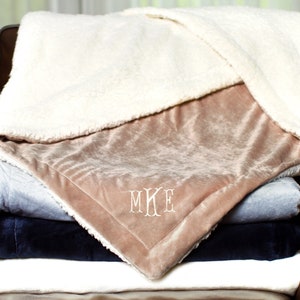 May include: A stack of four plush blankets in various colors, including white, gray, blue, and beige. The top blanket is beige and has the monogram "MKE" embroidered on it.