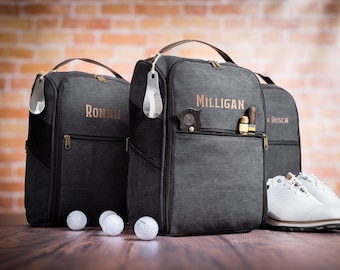 May include: Three black canvas golf shoe bags with personalized names, "Ronan", "Milligan", and "Bosch". The bags have a zippered pocket and a shoe horn attached. Three white golf balls are in front of the bags. A pair of white golf shoes are in front of the bag with the name "Bosch".