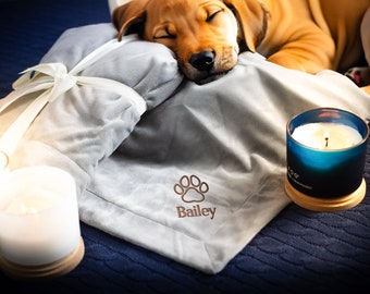 May include: A gray and white dog bed with a paw print and the name "Bailey" embroidered on it. A dog is sleeping on the bed. There are two candles on the table next to the bed.