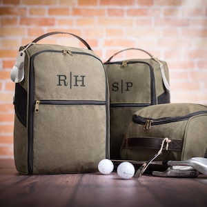 May include: Three green canvas golf shoe bags with black trim and leather accents. The bags have zipper closures and are monogrammed with the initials "R|H", "S|P", and a third bag with no visible initials. The bags are sitting on a wooden surface with golf balls and clubs.