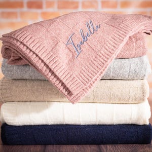May include: A stack of five knitted blankets in varying colors: dusty rose, gray, beige, white, and navy blue. The top blanket is embroidered with the name "Isabella" in a cursive font. The blankets are neatly folded and stacked on top of each other.